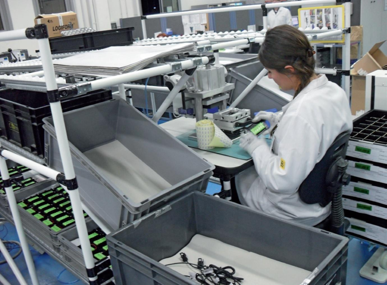 L'immagine raffigura un operatore che lavora in una linea di assemblaggio industriale, utilizzando un sistema di stoccaggio FIFO per gestire materiali e componenti. L'ambiente ordinato e l'utilizzo di scaffali e contenitori evidenziano un processo produttivo efficiente, ideale per la gestione di componenti elettronici o meccanici.
